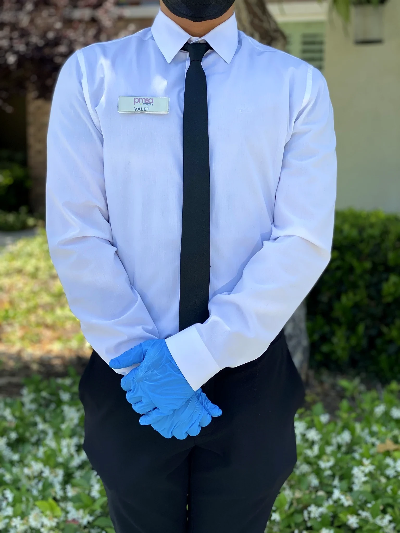 PMSA attendant in formal white dress shirt, black tie, valet badge, and blue gloves — hands clasped