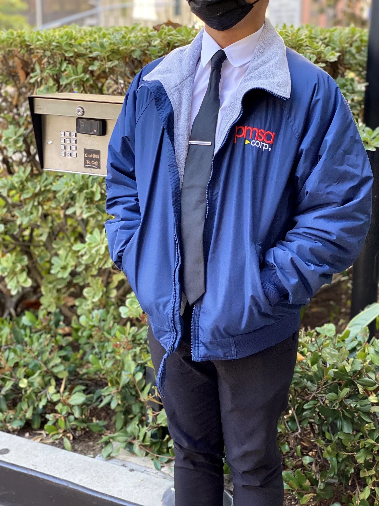 PMSA attendant in branded blue jacket, white shirt, tie, and mask standing at a building entry keypad